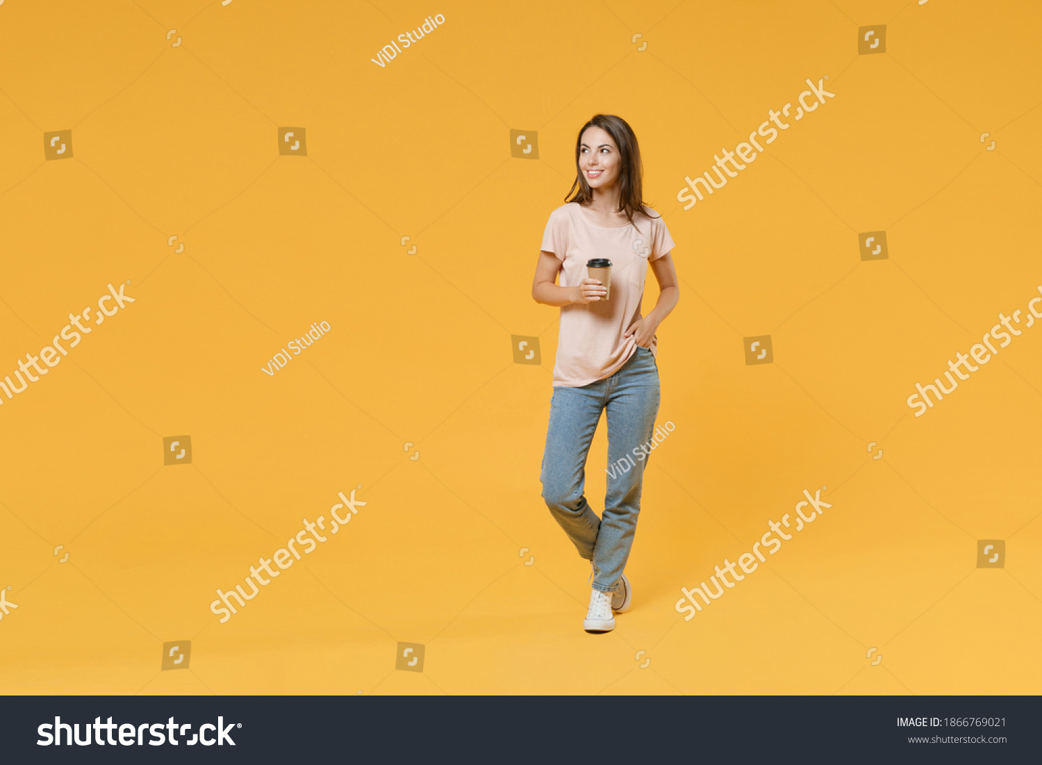 Full length portrait of smiling young brunette woman 20s wearing pastel pink casual t-shirt posing hold paper cup of coffee or tea looking aside isolated on bright yellow color wall background studio #1866769021