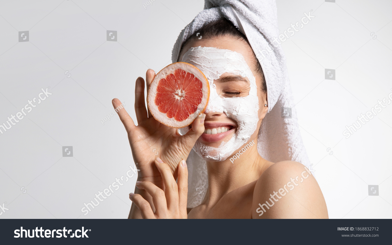Close up of happy young playful teen girl in moisturizing mask and towel holds grapefruit covers eye. Advertising poster of facial eco-friendly skincare products. Morning beauty routine #1868832712