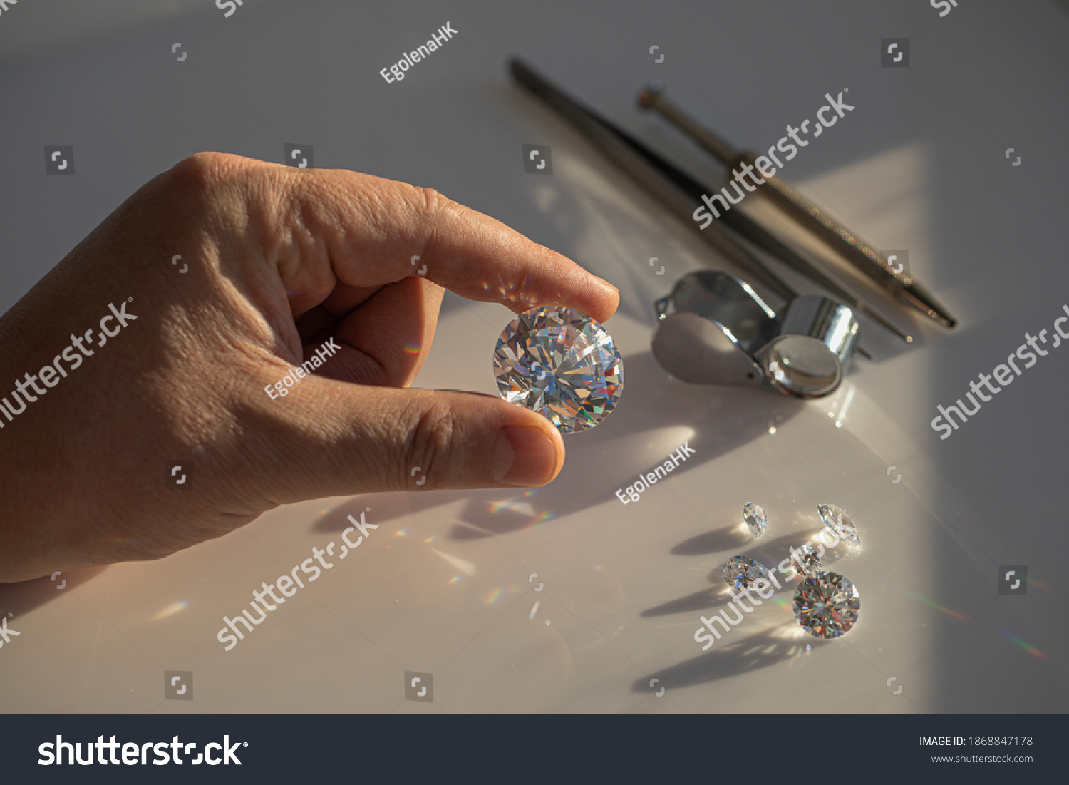 Close-up of the hand of jeweller gemologist with large size round cut diamond on desktop with magnifier and tweezers. Buyer diamond expert checking color of diamond in natural light. #1868847178
