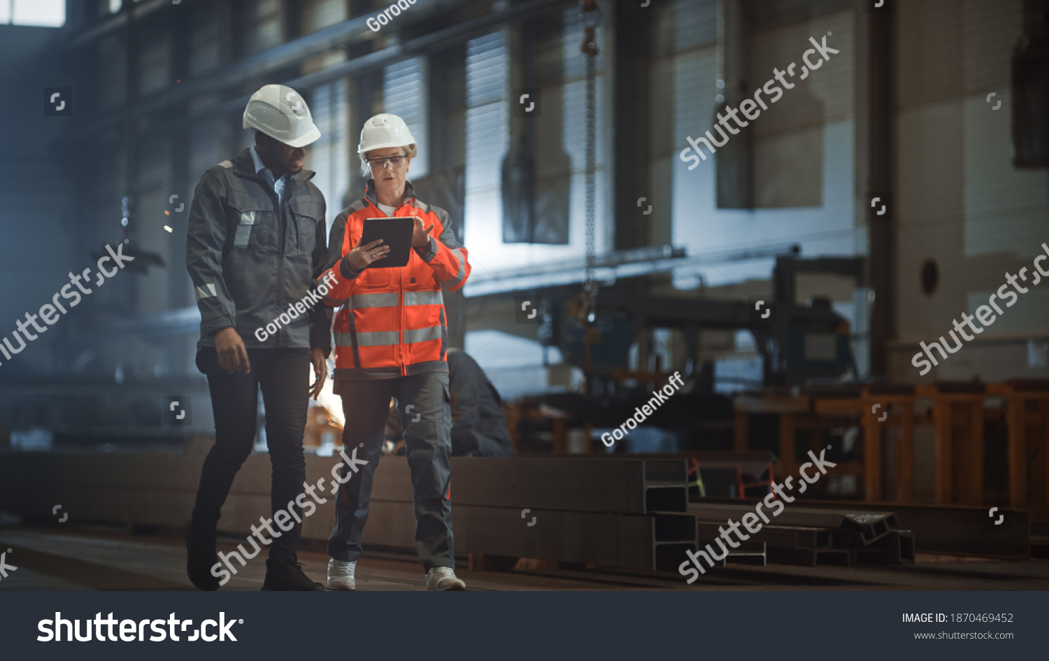 Two Heavy Industry Engineers Walk in Steel Factory, Use Tablet and Discuss Work. Industrial Worker Uses Angle Grinder in the Background. Black African American Specialist Talks to Female Technician. #1870469452