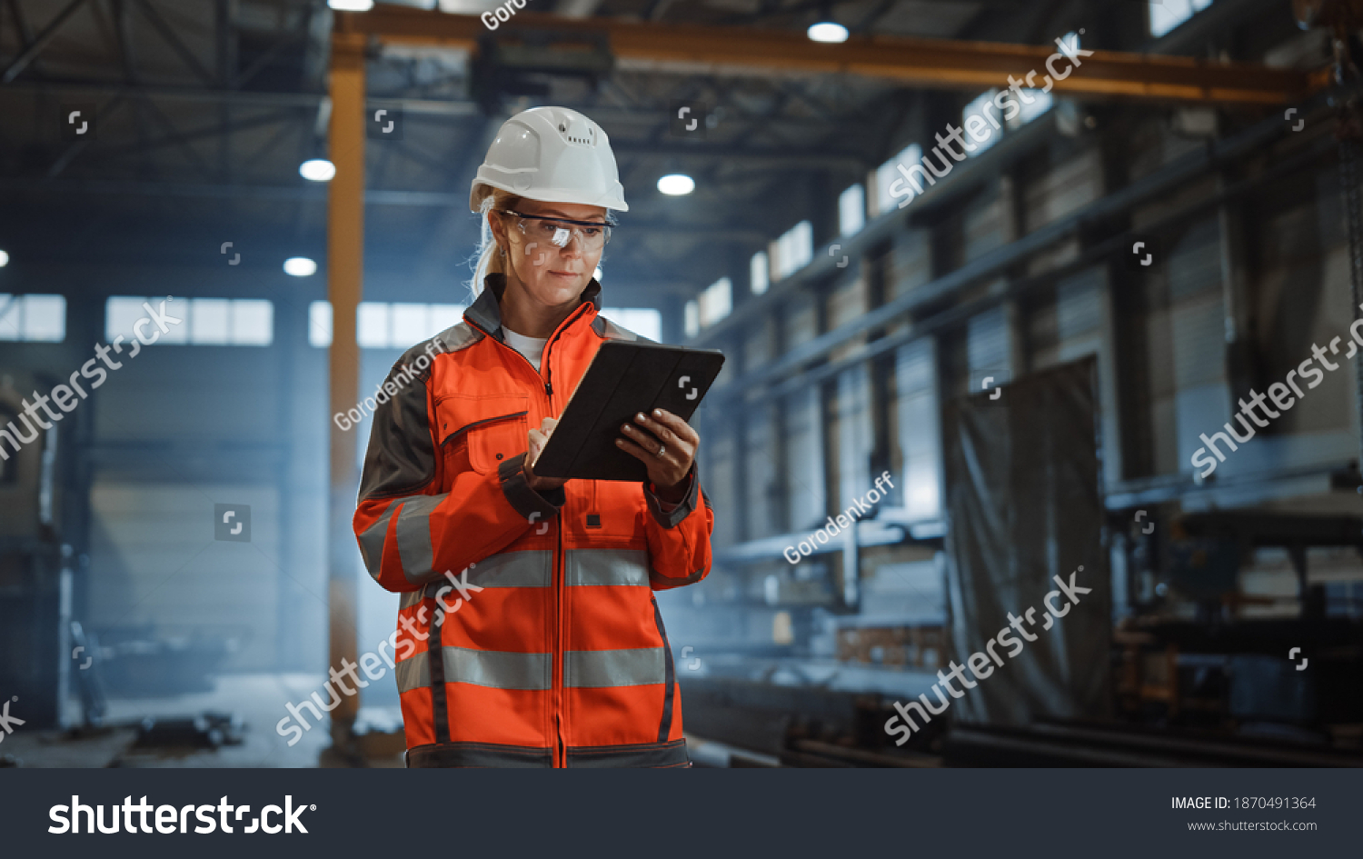 Professional Heavy Industry Engineer Worker Wearing Safety Uniform and Hard Hat, Using Tablet Computer. Serious Successful Female Industrial Specialist Walking in a Metal Manufacture Warehouse. #1870491364