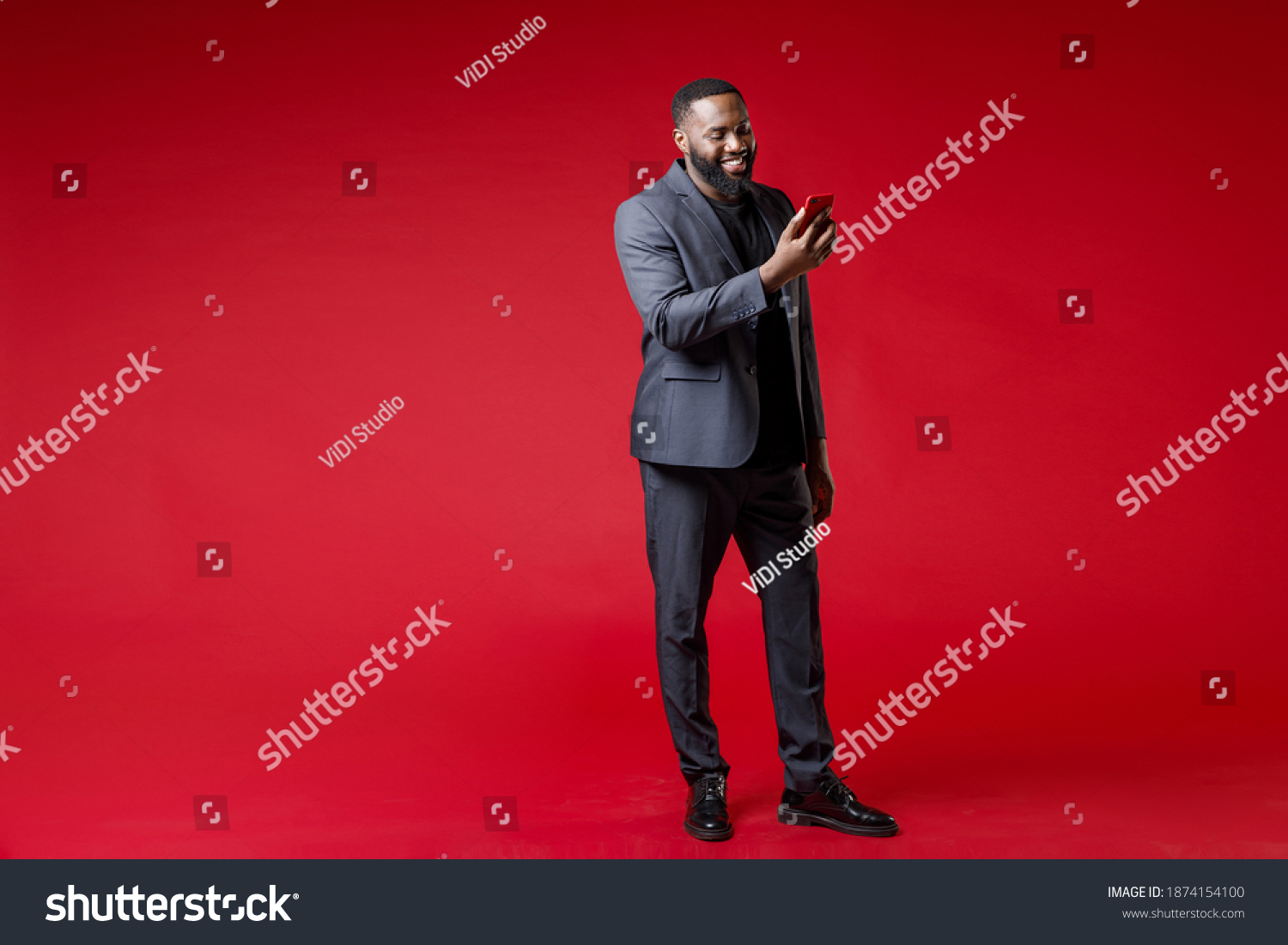 Full length of smiling successful young african american business man 20s in classic jacket suit using mobile cell phone typing sms message isolated on bright red color background studio portrait #1874154100
