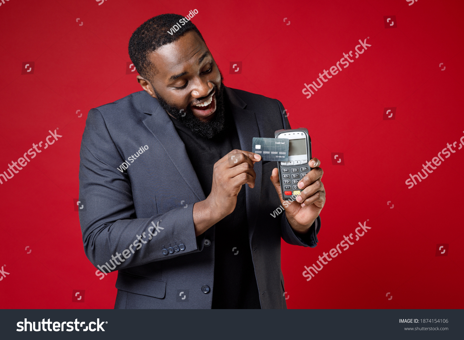 Cheerful young african american business man 20s in classic jacket suit hold wireless modern bank payment terminal to process acquire credit card payments isolated on red background studio portrait #1874154106