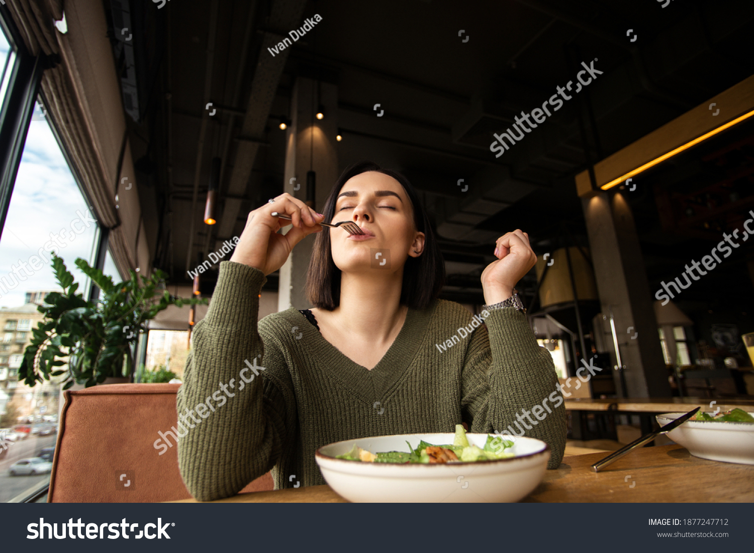 Satisfied young woman enjoys tasty salad in a restaurant, putting fork into her mouth and closing her eyes. Good customer service. Healthy diet concept. Weight loss without struggle. #1877247712