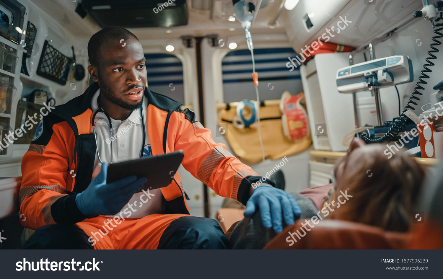 Black African American EMS Professional Paramedic Using Tablet Computer to Fill a Questionnaire for the Injured Patient on the Way to Hospital. Emergency Medical Care Assistant Works in an Ambulance. #1877996239