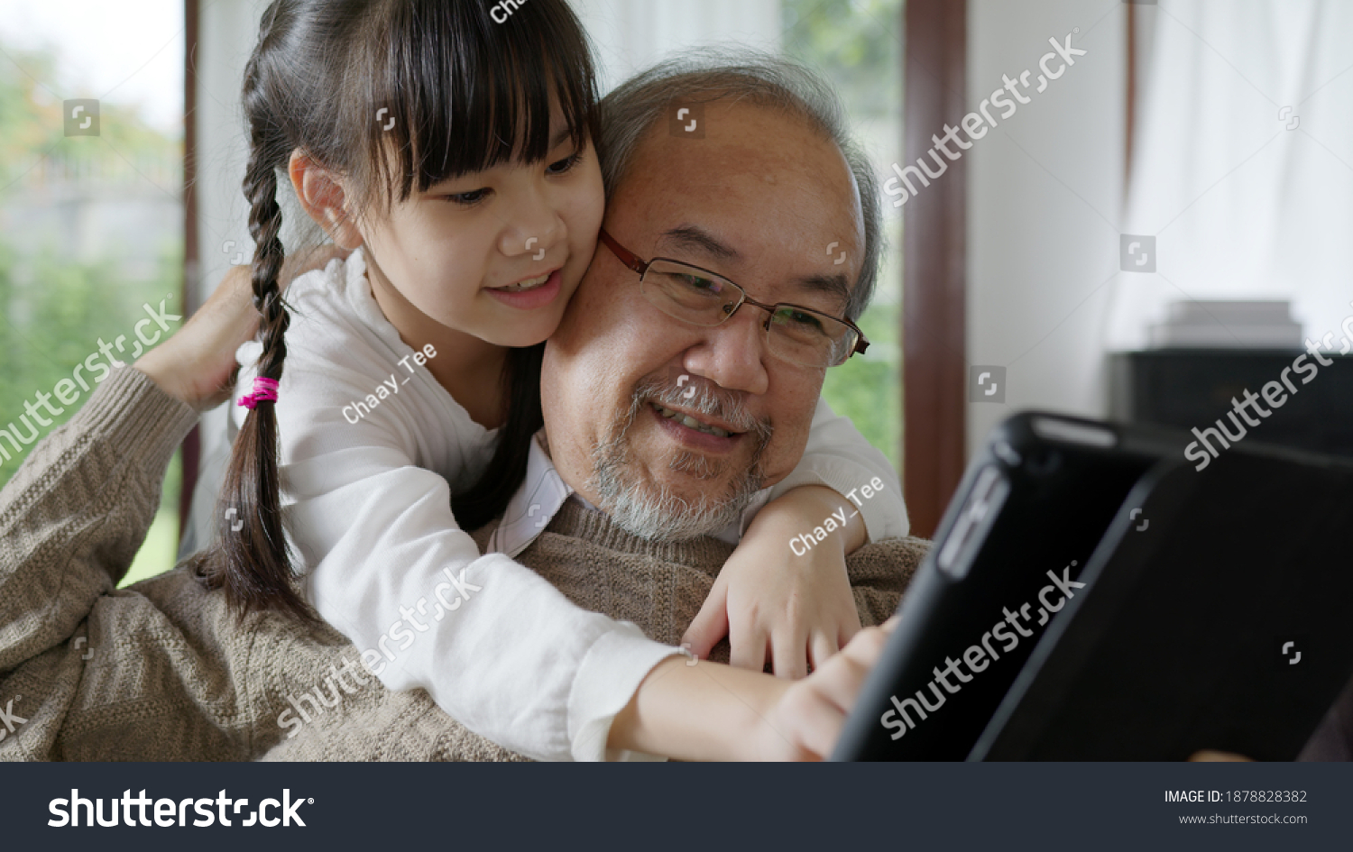 Candid of old senior asian grandparent play and watch with kid grandchildren with technology on computer tablet at home in bonding relationship in family. Young girl hug older man from back. #1878828382