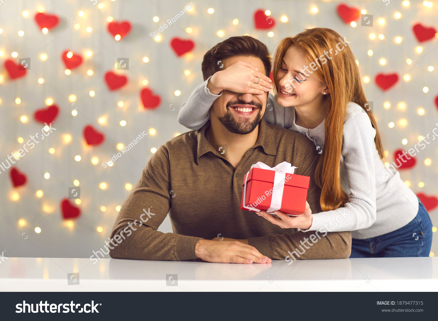 Young couple in love celebrating Saint Valentine's Day or relationship anniversary. Happy woman covering boyfriend's eyes giving him surprise gift. Smiling man getting present from loving girlfriend #1879477315