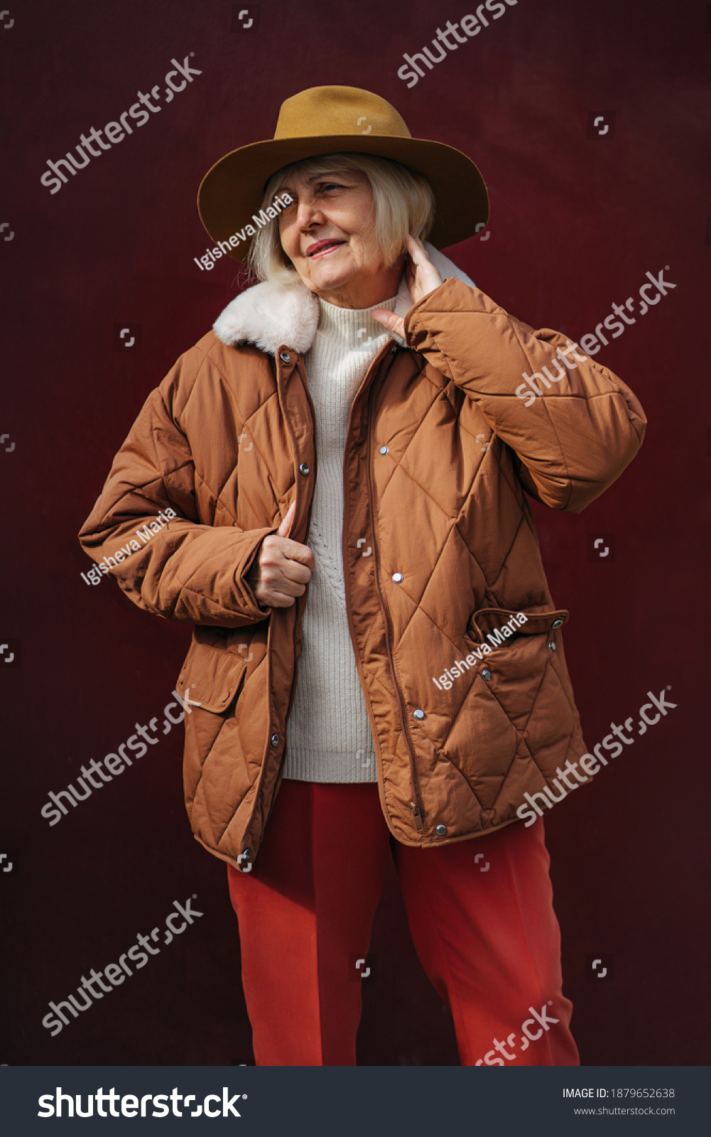 Senior woman in trendy outerwear and hat looking away while standing against vinous wall. Aged female in stylish outerwear #1879652638