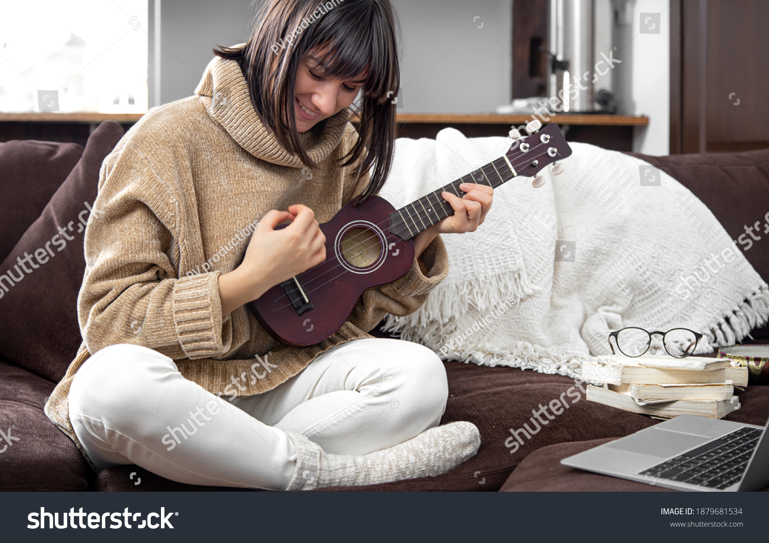 Young cheerful girl in a sweater learns to play the ukulele. The concept of online learning, home education. #1879681534