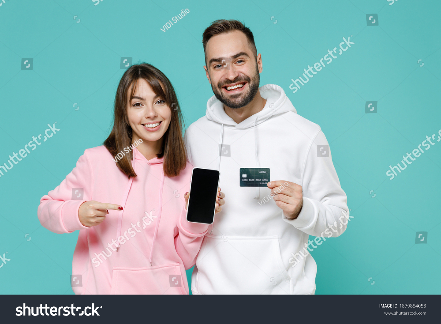 Smiling young couple two friends man woman 20s wearing white pink hoodie pointing on mobile phone with blank empty screen credit bank card isolated on blue turquoise colour background studio portrait #1879854058