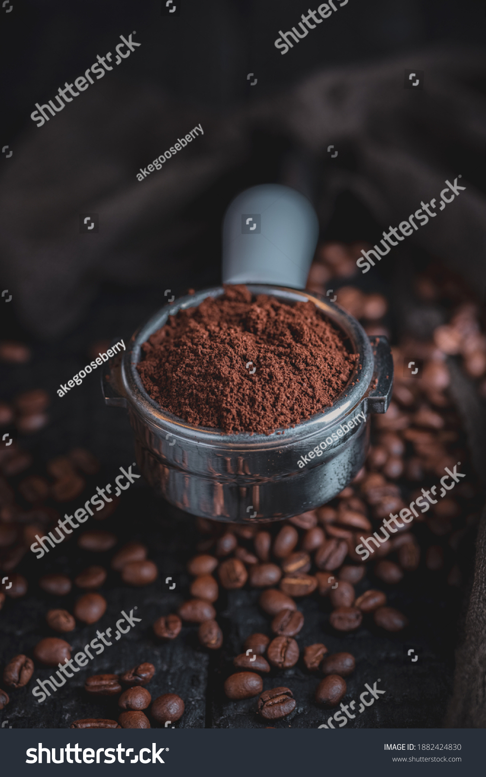 Tamped puck of coffee grounds within basket of portafilter and coffee beans spilled around in a dark and moody scene of natural light. #1882424830