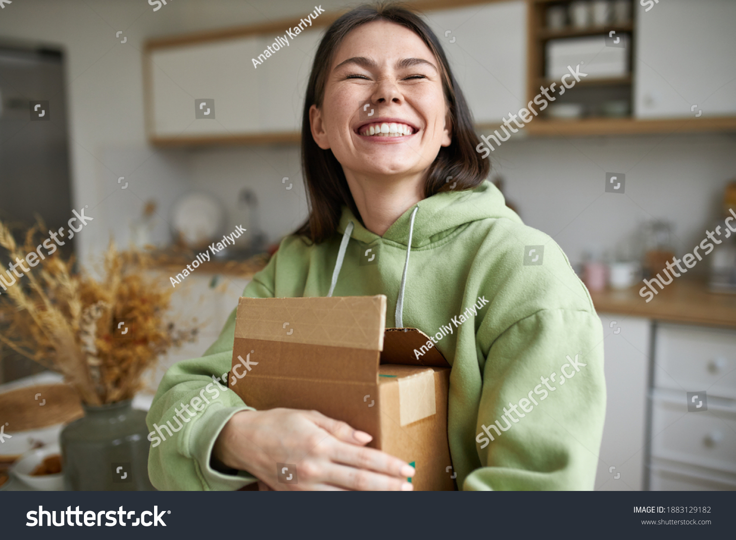 Cheerful teenage girl expressing positive emotions receiving unexpected birthday gift holding parcel and smiling with pleasure. Dark haired young woman posing with cardboard box with new cosmetics #1883129182