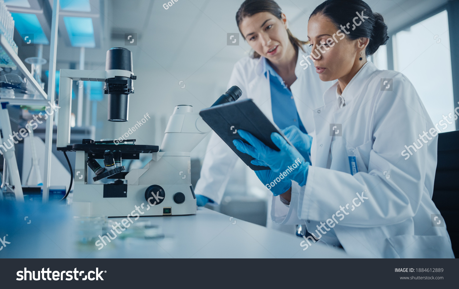 Modern Medical Research Laboratory: Two Female Scientists Working, Using Digital Tablet, Analysing Samples, Talking. Advanced Scientific Pharmaceutical Lab for Medicine, Biotechnology Development #1884612889