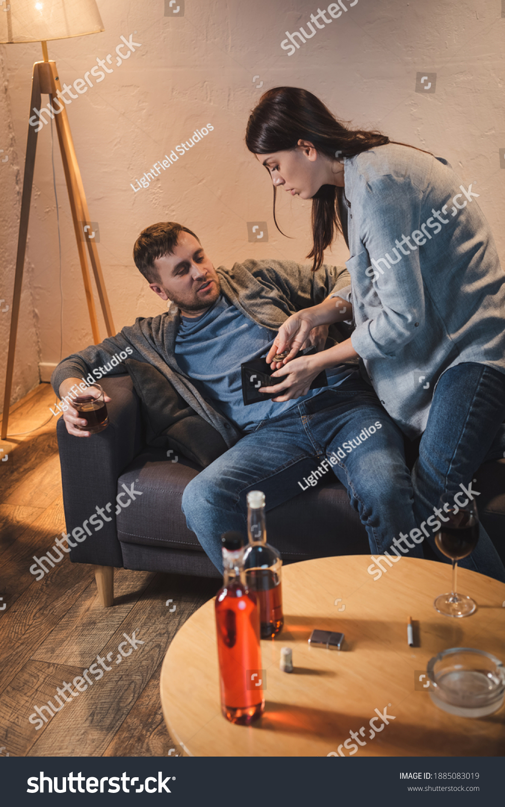 woman showing coins and empty wallet to drunk husband sitting on sofa with glass of whiskey #1885083019