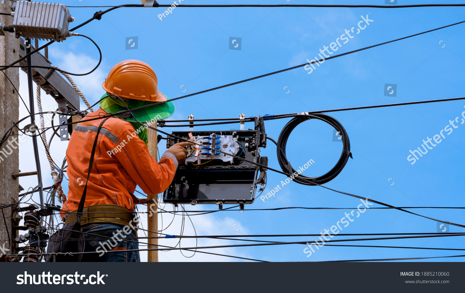 Rear view of technician on wooden ladder checking fiber optic cables in internet splitter box on electric pole against blue sky background #1885210060
