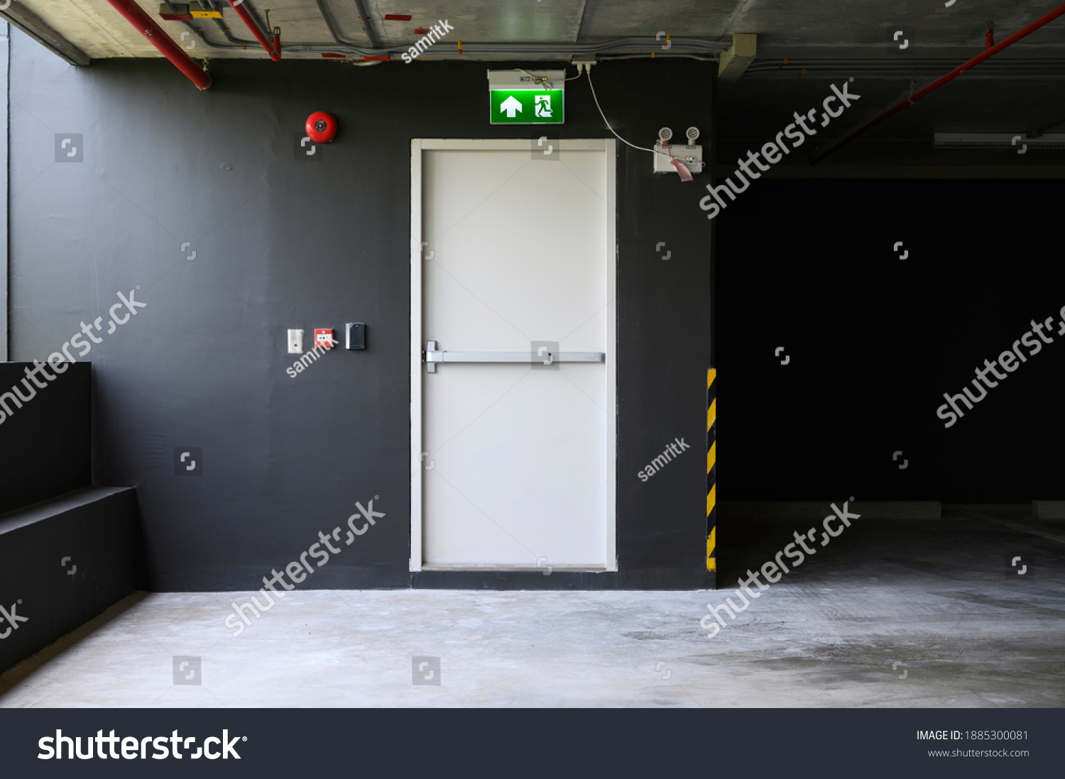 A fire exit with the sign and red alarm bell above the white door on the parking building. #1885300081