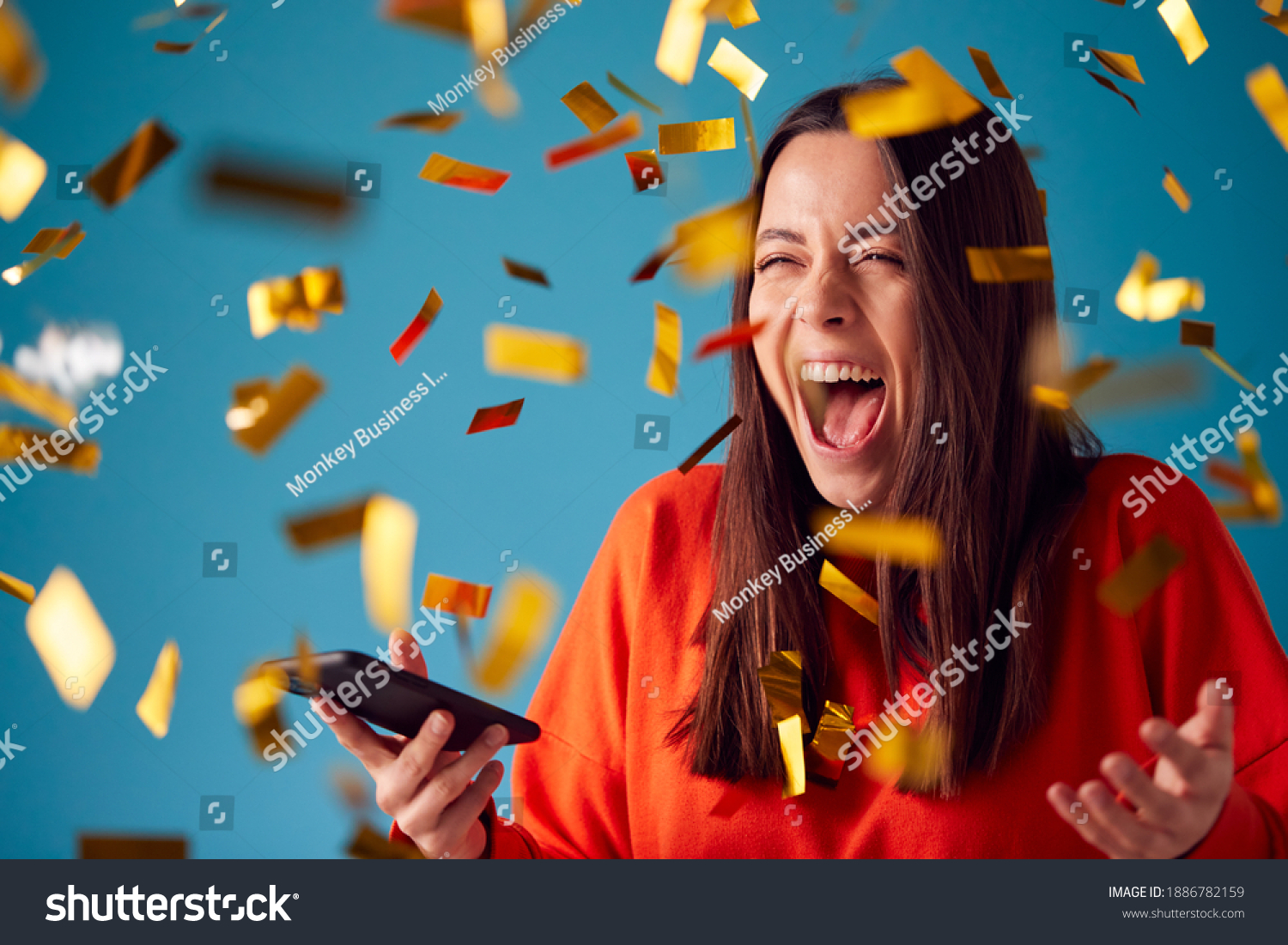 Celebrating Young Woman With Mobile Phone Winning Prize And Showered With Gold Confetti In Studio #1886782159