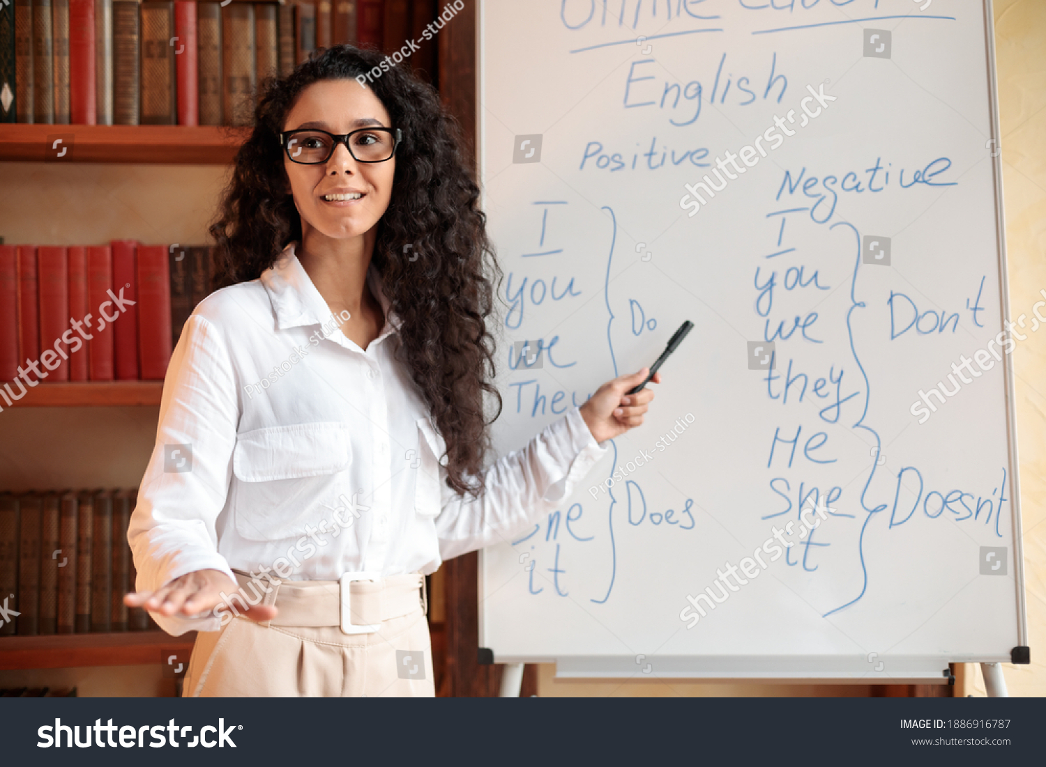 Foreign Language. Portrait of confident smiling young female lecturer wearing glasses teaching English pointing at grammar rules on board with marker, looking back at students at classroom #1886916787