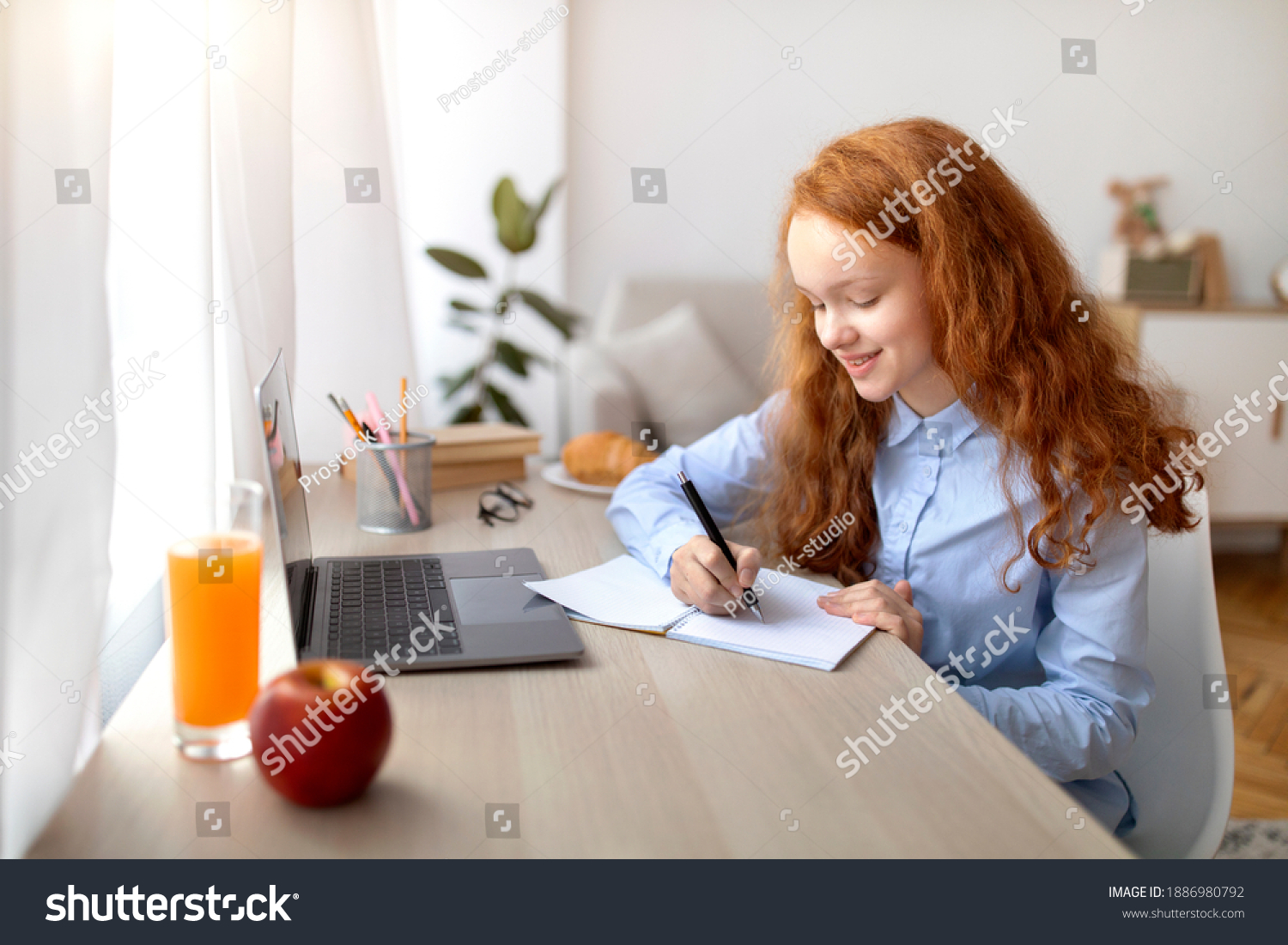 Remote Education Concept. Smiling young teenage girl sitting at table, using laptop and writing in her notebook or diary. Schoolgirl watching online course, taking notes, doing homework #1886980792