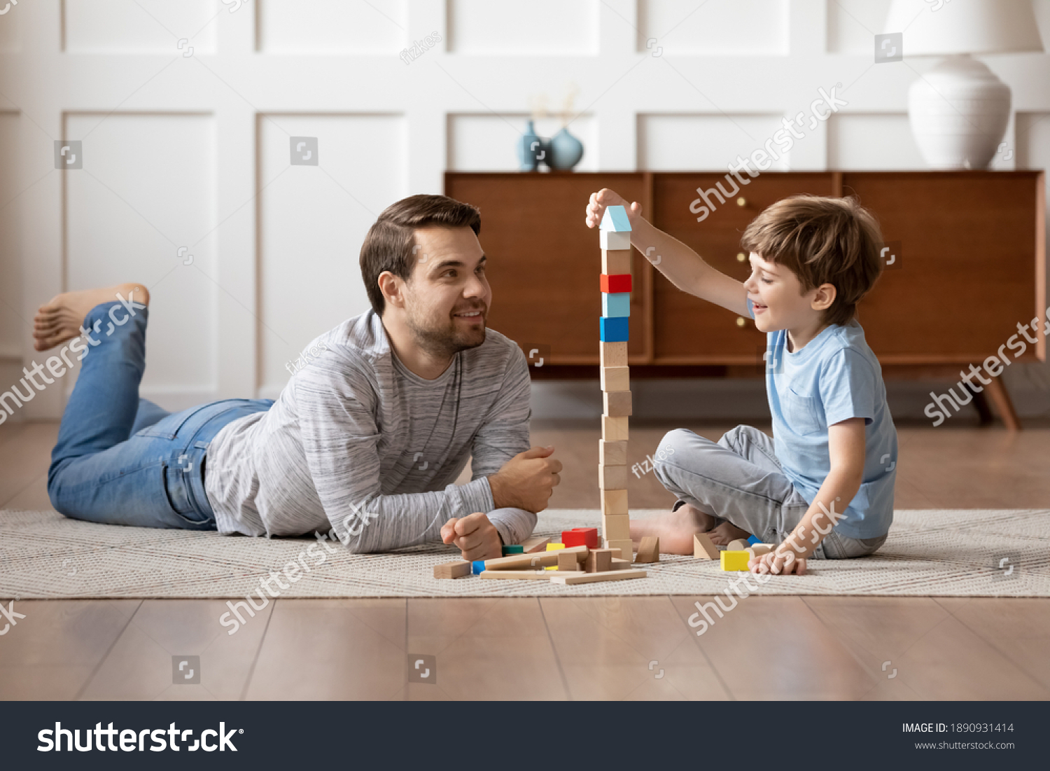 Caring young Caucasian father and small son sit on warm floor at home engaged in funny game together. Loving dad and little boy child have fun play build construct with wooden blocks bricks. #1890931414