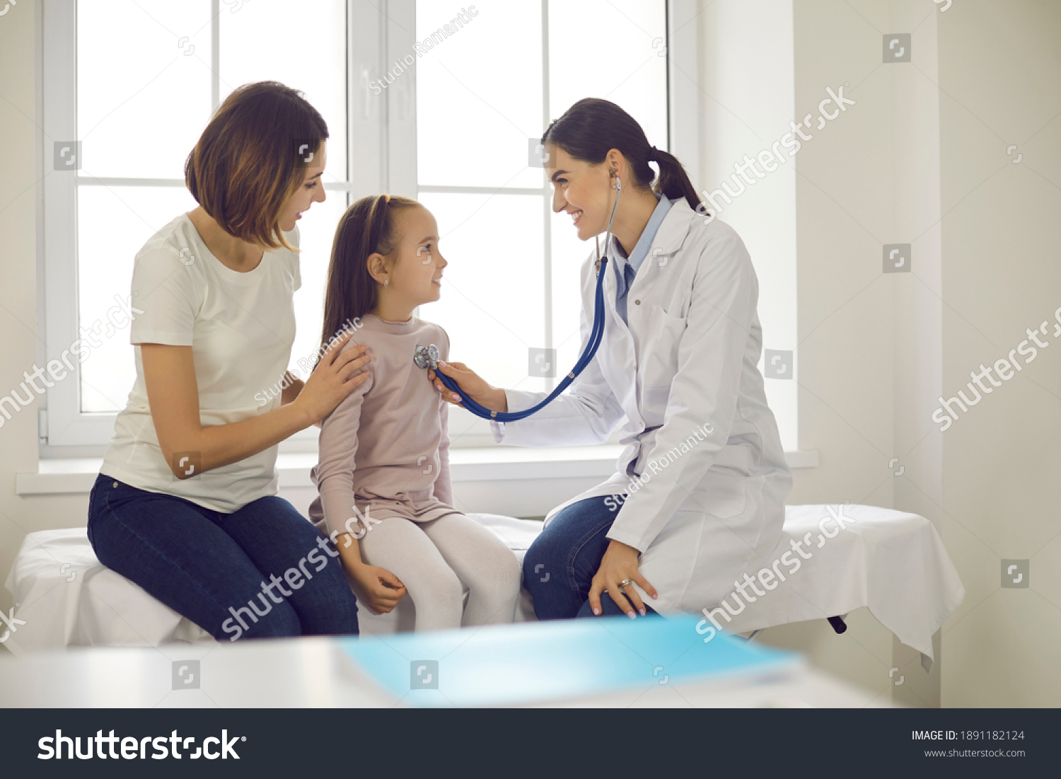 Mother and child seeing family practitioner. Smiling pediatrician with stethoscope checking little girl's lungs. Friendly doctor listening to young patient's breath or heartbeat during medical exam #1891182124