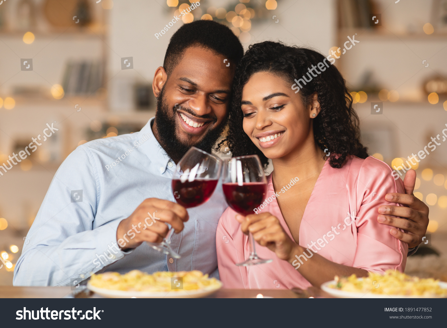 Cheers. Beautiful lovely young african american couple sitting at table in restaurant or at home, drinking red wine, holding glasses during romantic dinner. Smiling black man embracing his happy woman #1891477852