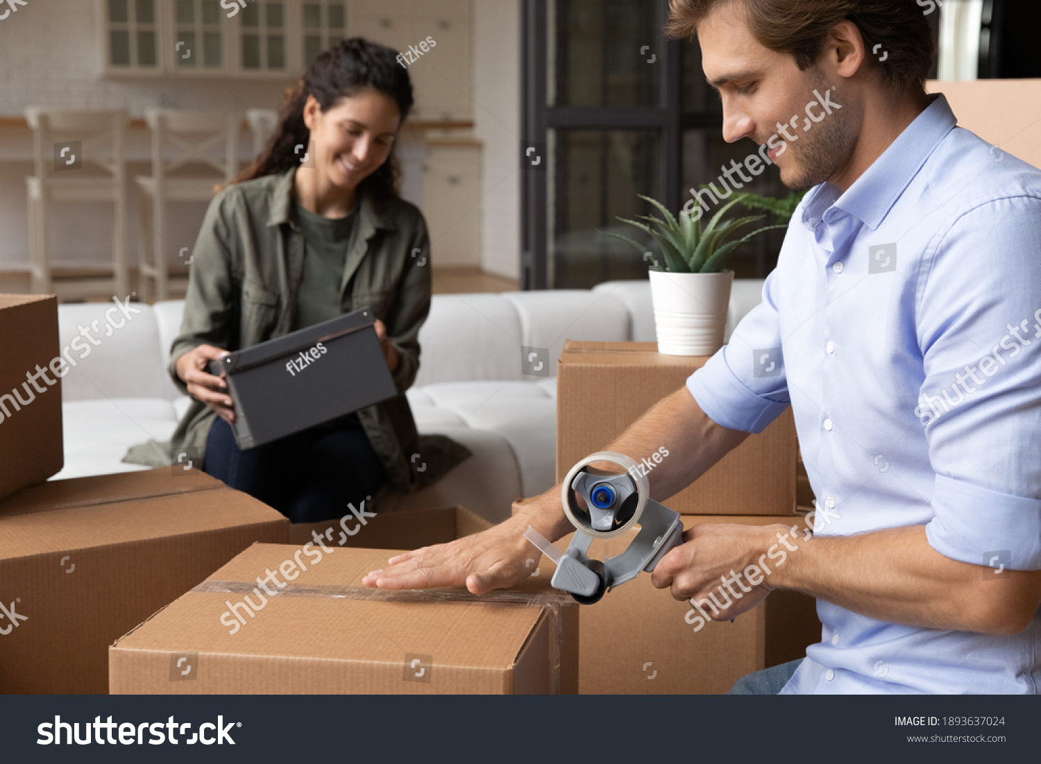 Happy young Caucasian man and woman seal wrap packages with tape dispenser relocate to new house. Smiling millennial couple renters pack boxes with adhesive scotch, moving to own home together. #1893637024