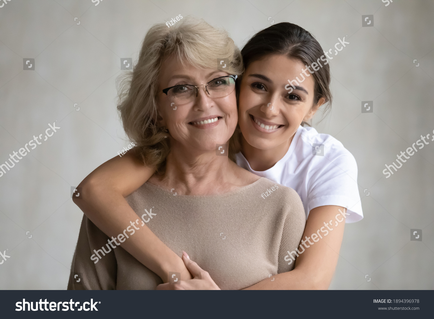 Head shot portrait loving grown up daughter hugging middle aged mother from back, looking at camera, happy mature grandmother and granddaughter posing for family photo on grey background together #1894396978
