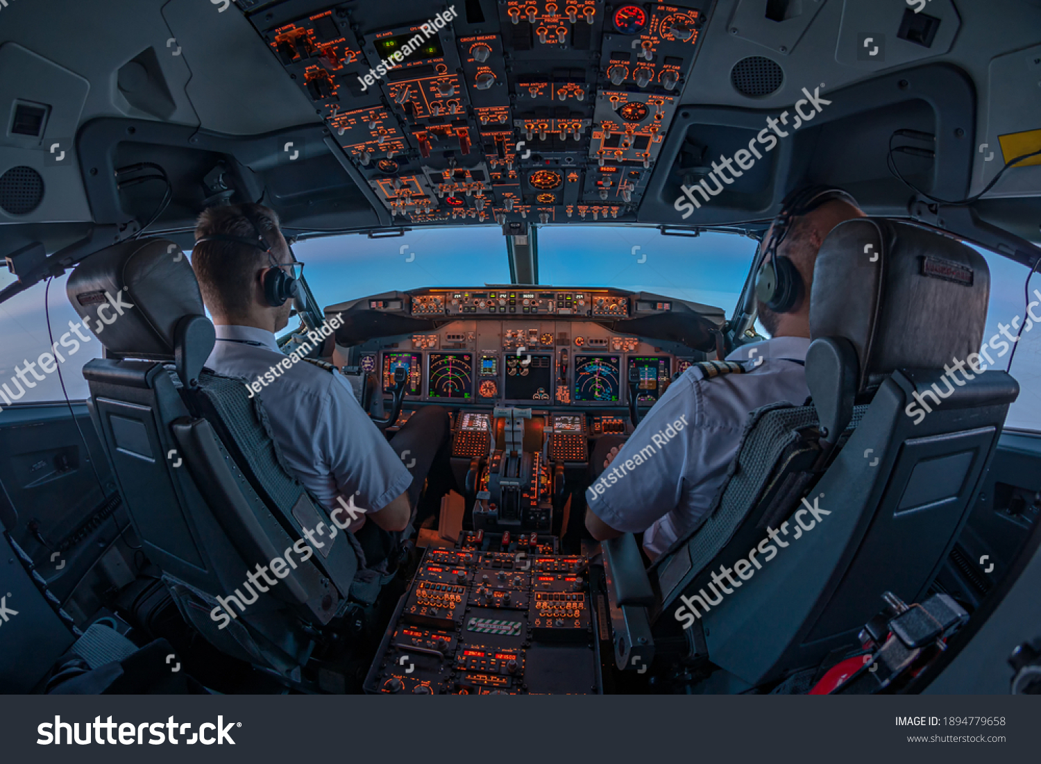 Pilots in the cockpit of jet commercial airplane during the flight with first rays of the warm sunrise entering through the flight deck window #1894779658