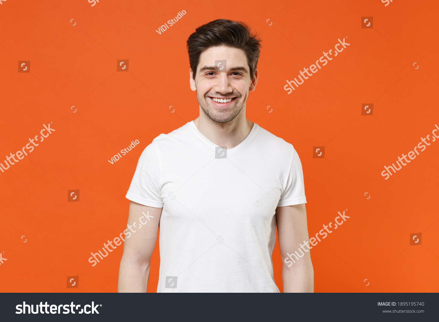 Smiling cheerful funny handsome attractive young man 20s wearing basic casual white blank empty t-shirt standing looking camera isolated on bright orange colour wall background, studio portrait #1895195740