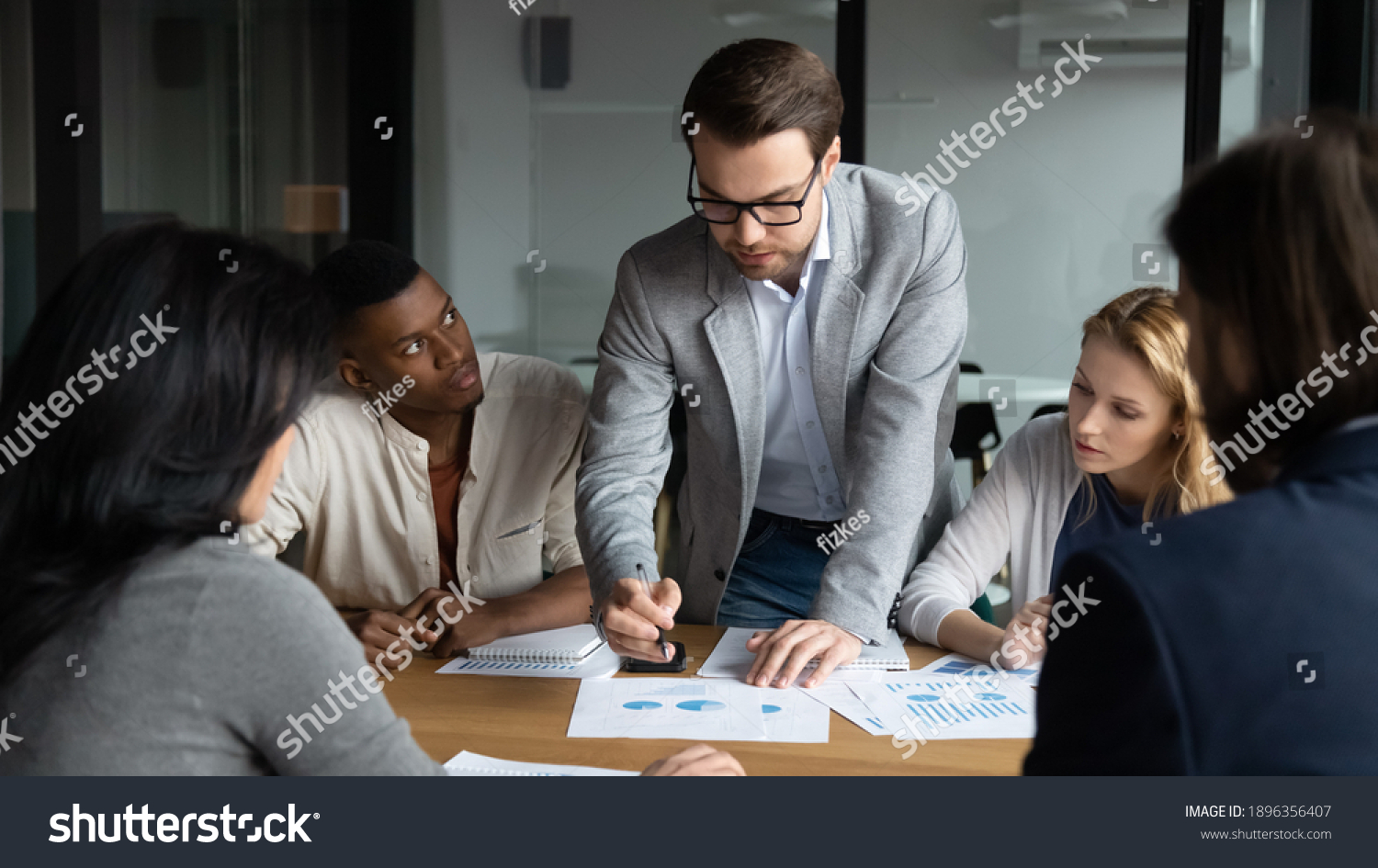 Skilled focused young businessman boss leader in eyeglasses analyzing sales data statistics or explaining marketing strategy to motivated diverse multiracial colleagues at brainstorming meeting. #1896356407