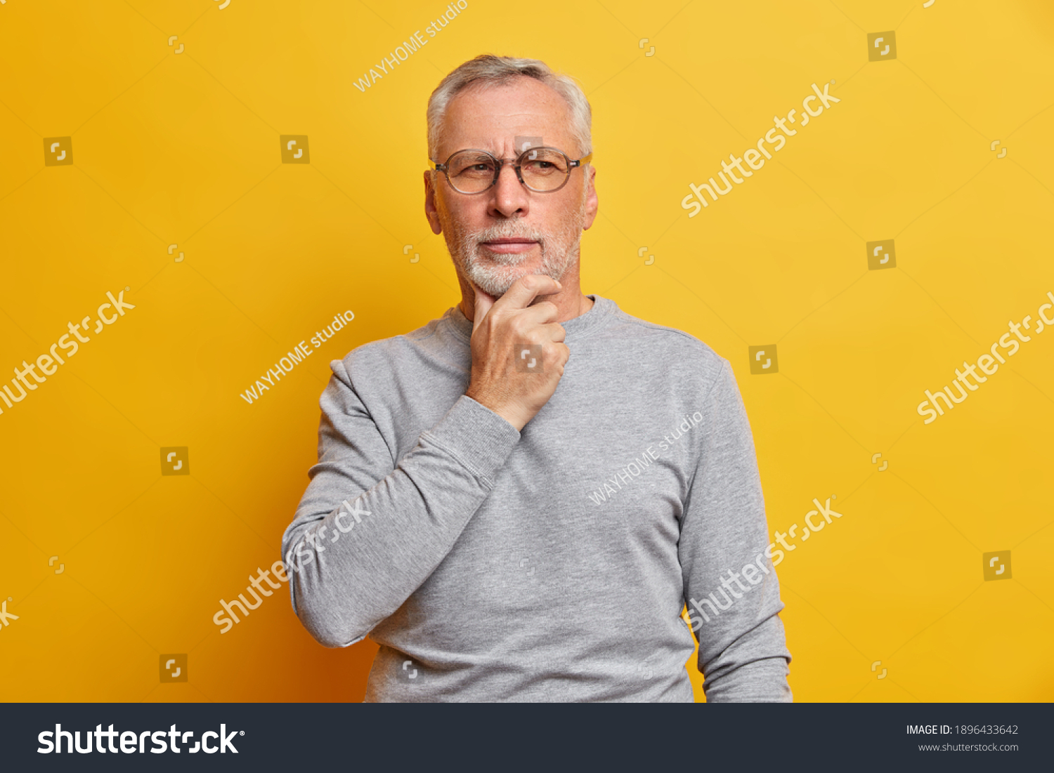 Senior thougthful man holds chin and looks pensively aside makes plannings wears spectacles and casual grey jumper isolated over vivid yellow background. Handsome grandfather considers something #1896433642