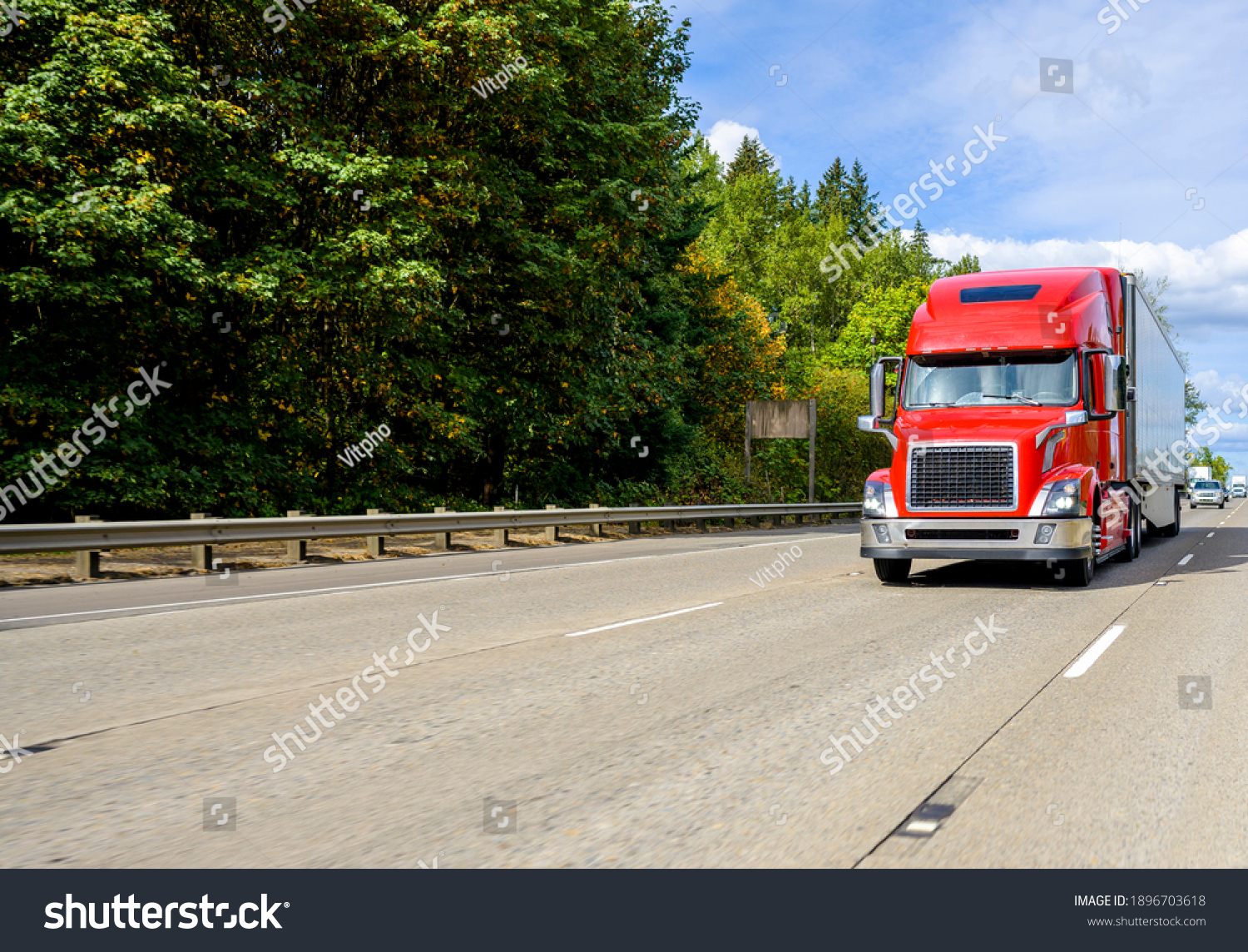 Red big rig industrial grade bonnet long hauler diesel semi truck with high roof cab and refrigerator semi trailer running with commercial cargo on the wide highway road with green trees hillside #1896703618