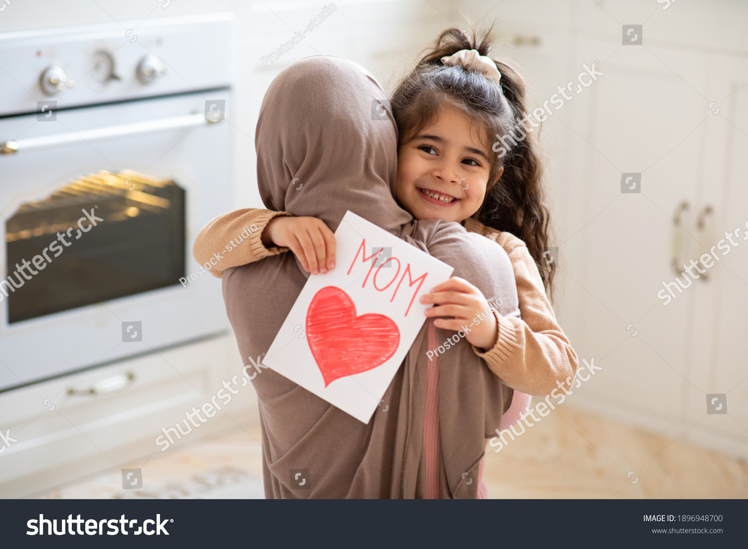 Smiling little girl holding greeting card for Happy Mother's Day with drawn red heart and hugging her muslim mom. Loving islamic family bonding together at home, closeup shot with free space #1896948700