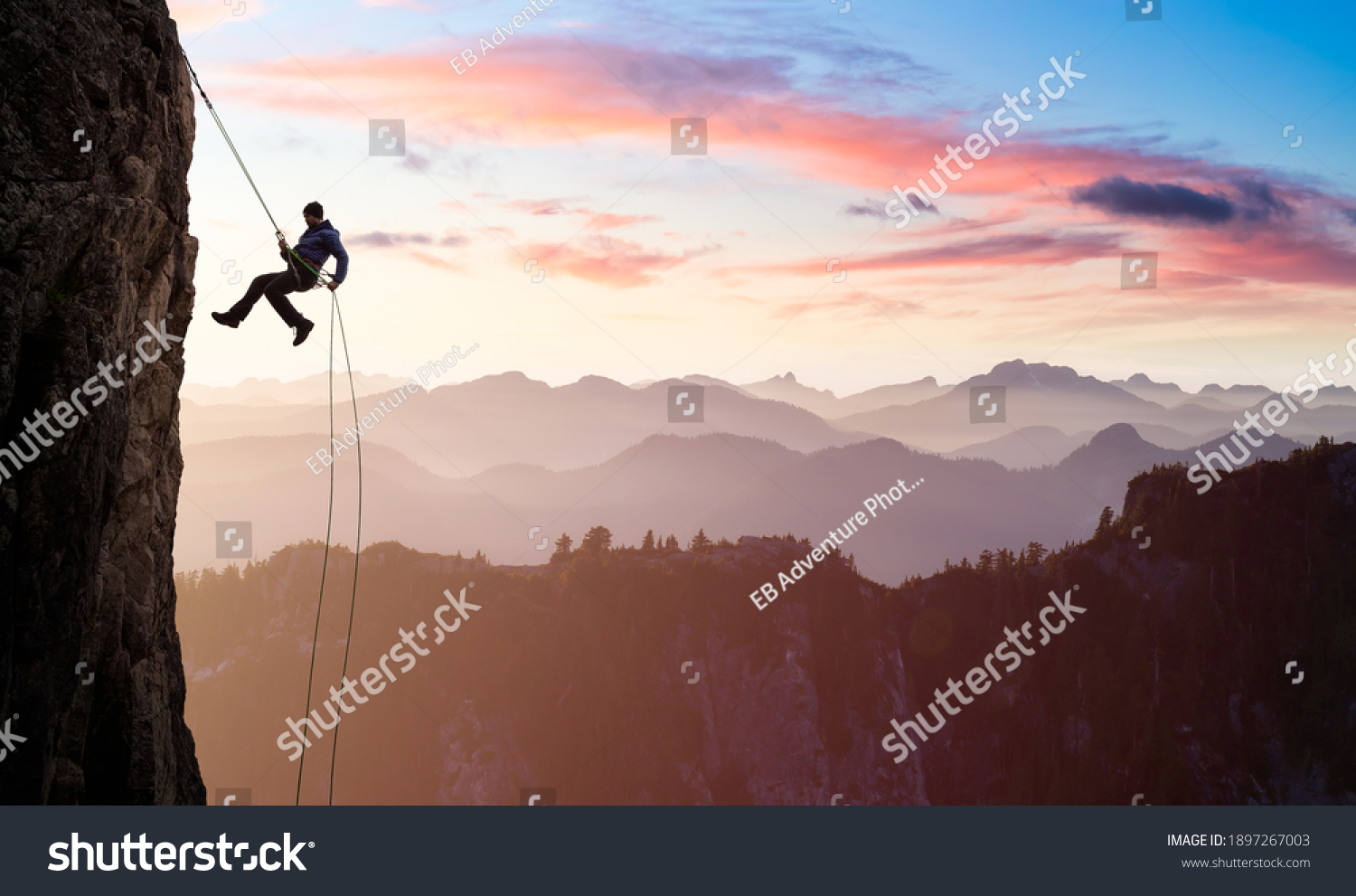 Adventurous Man Rappelling from Cliff. Aerial view of the mountains during a colorful and vibrant sunset or sunrise. Landscape taken in British Columbia, Canada. composite. Concept: Adventure #1897267003