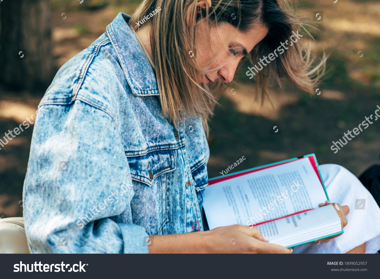 Closeup of a beautiful blonde female reads a book while sitting on the bench on a sunny day in the street. A pretty young woman in a jeans jacket takes a rest outside and learning from a book. #1899052957