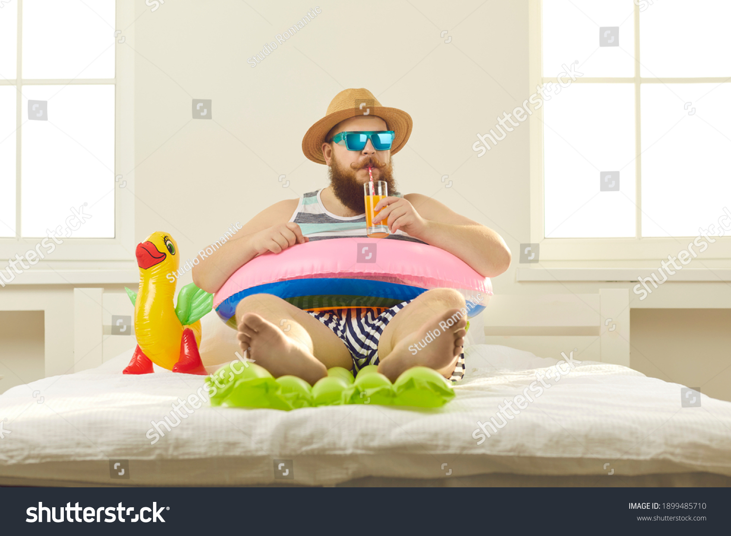 Funny young man in sunglasses and summer wear sipping beach cocktail, sitting barefoot in inflatable lifebuoy at home. Covid-19 quarantine, vacation in lockdown, canceled holiday travel plans concept #1899485710