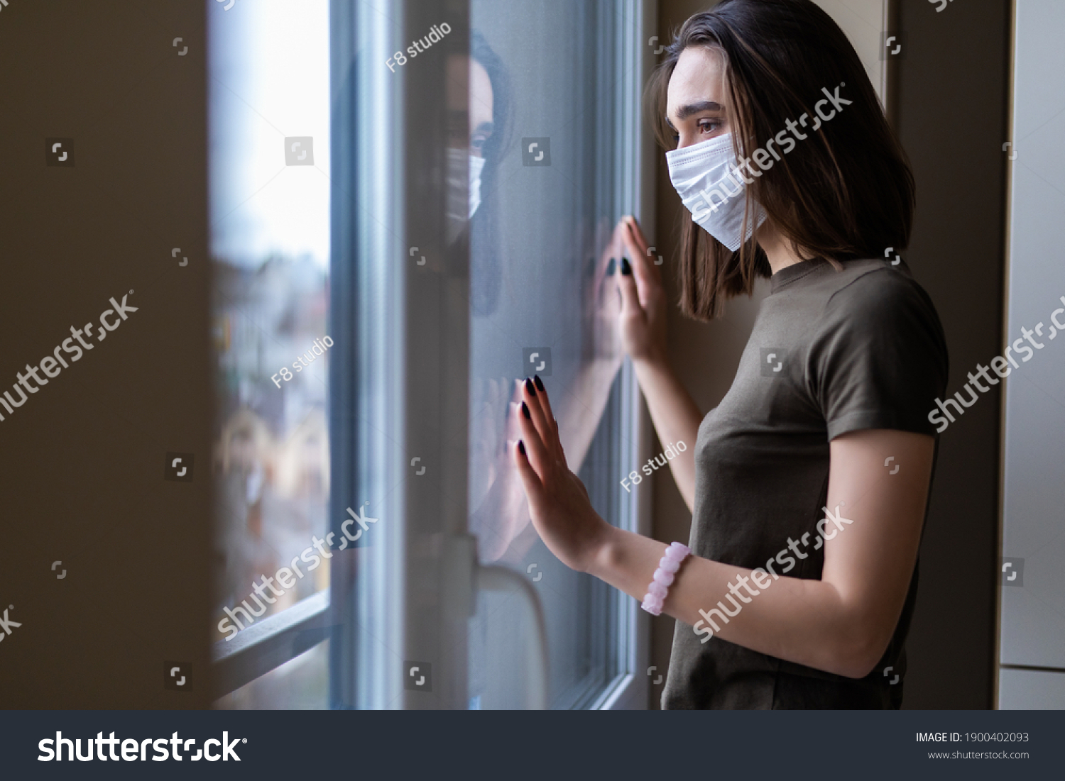 Sad young woman in a medical mask looks out of the window. Quarantine during the coronavirus pandemic. #1900402093