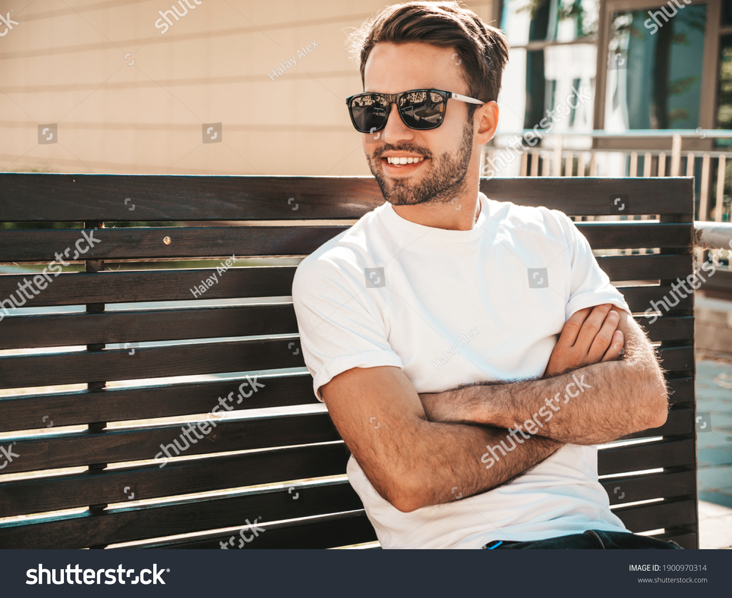 Portrait of handsome smiling stylish hipster lambersexual model.Man dressed in white T-shirt. Fashion male sitting at the bench in the street background in sunglasses. Crossed arms #1900970314