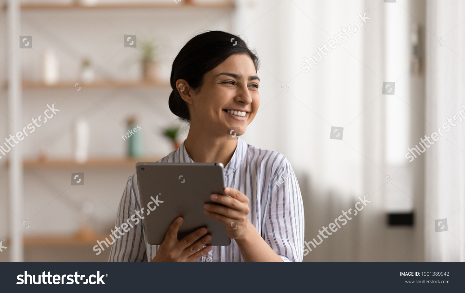 Full of dreams. Smiling mixed race lady web shop customer hold pad pc look aside choose goods plan purchases. Positive indian female relax at home with digital tablet enjoy modern device possibilities #1901389942