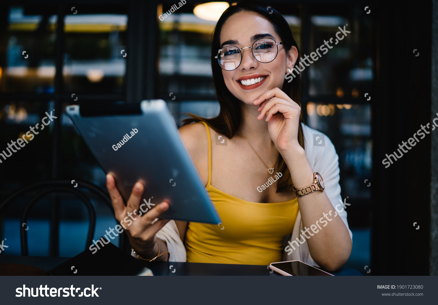 Portrait of cheerful Caucasian female blogger in eyewear smiling at camera during time for online networking via digital tablet, joyful hipster girl with modern touch pad posing in street cafe #1901723080