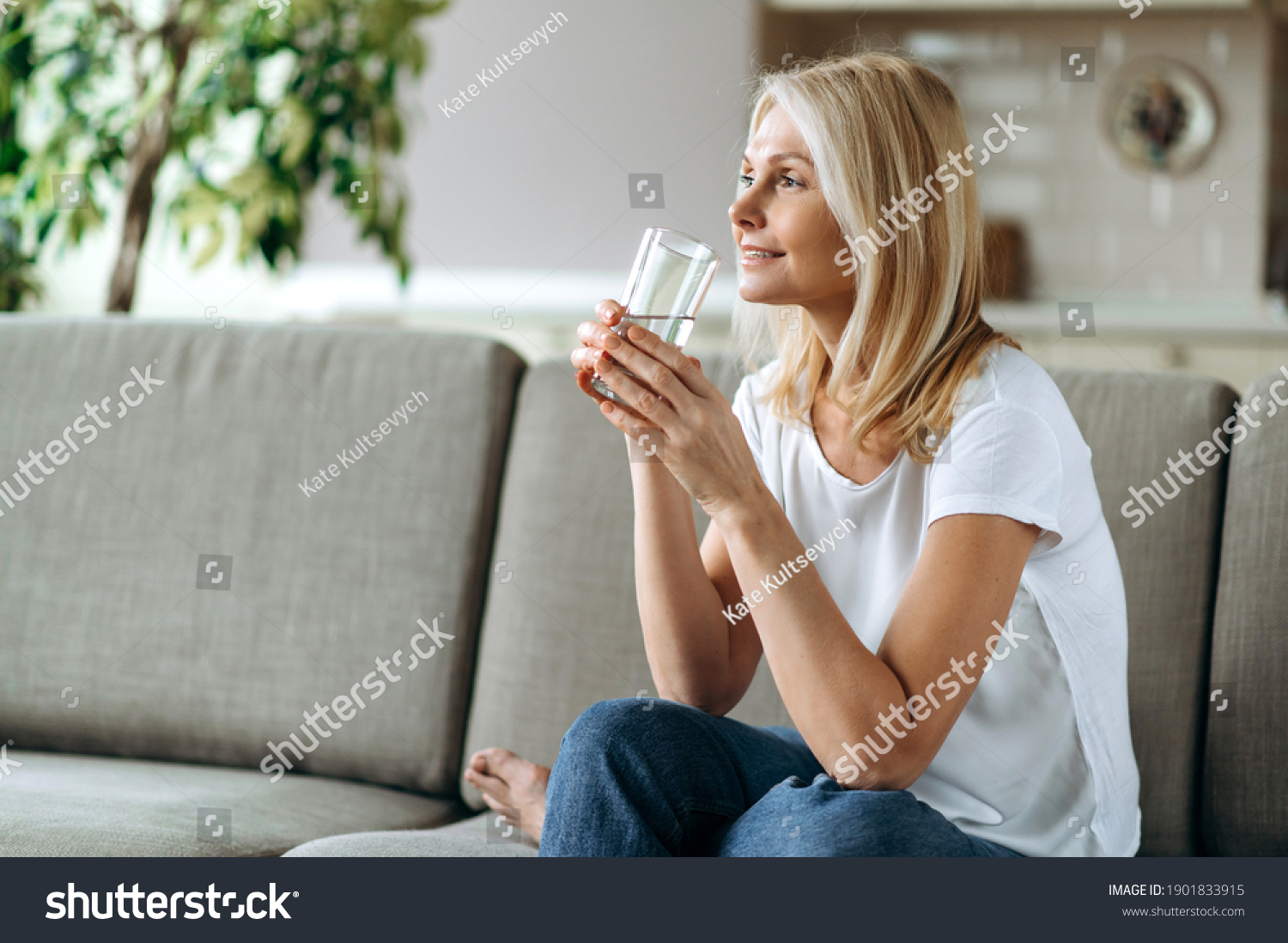 Healthcare concept. Mature caucasian blonde dressed in a casual wear sits on the couch in a living room, holding a glass of pure water in a hand and smiling, follow healthy lifestyle #1901833915