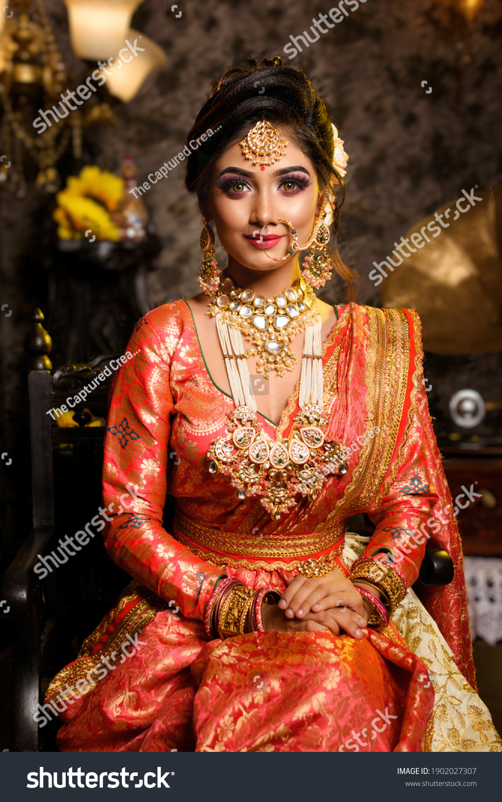 Magnificent young Indian bride in luxurious bridal costume with makeup and heavy jewellery sitting in a chair with classic vintage interior in studio lighting. Wedding fashion. #1902027307