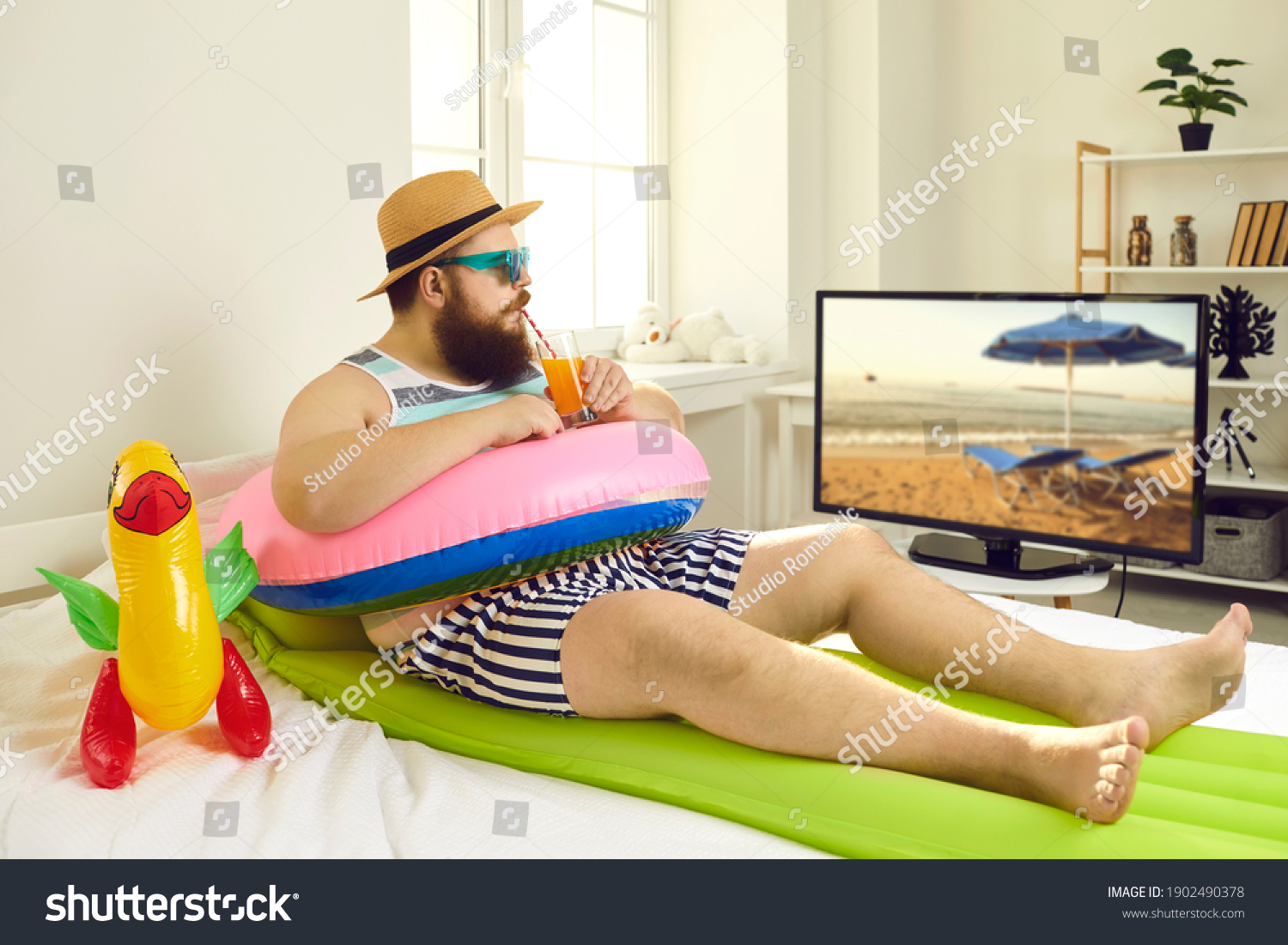 Funny young man with sunglasses and inflatable beach toys sipping cocktail and watching travel show on TV. Concept of canceled summer holiday plans, vacation in lockdown at home or Covid-19 quarantine #1902490378