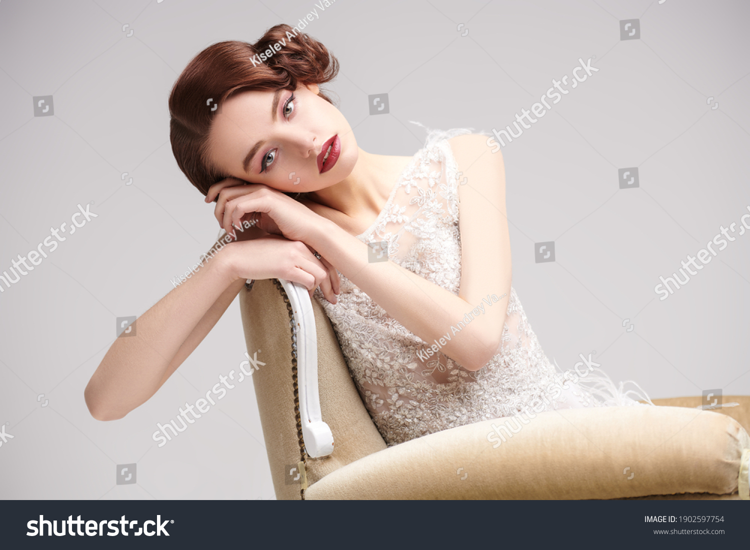 Portrait of a beautiful chic woman posing in a luxury white dress in vintage armchair. Evening makeup and hairstyle of the 20s.  #1902597754