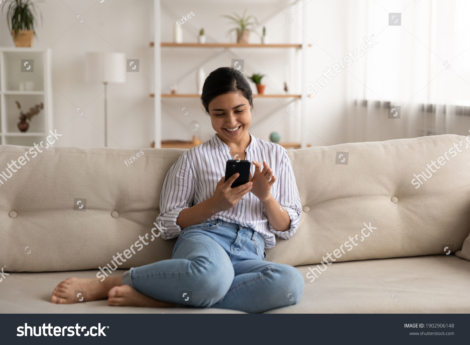 Happy indian lady relax at home alone sit on large couch in comfortable pose share good news at social media via cellphone. Smiling mixed race woman enjoy weekend order goods food online in phone app #1902906148