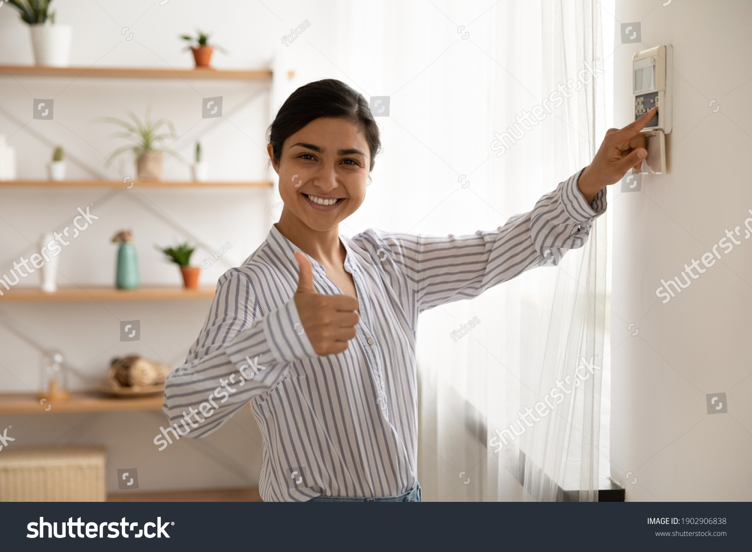 Portrait of smiling indian lady use domestic fire alarm system switch button on wall keypad show thumb up. Young female satisfied user of home security equipment look at camera trust recommend safety #1902906838