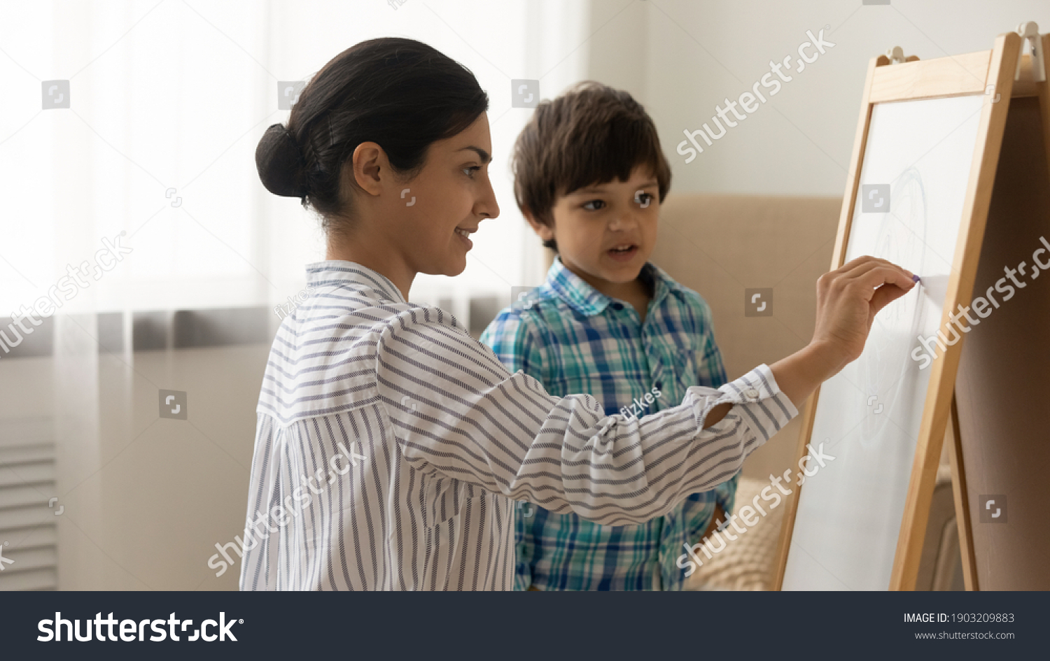 Attentive little indian boy preschooler take painting class from young millennial woman art teacher. Interested small child son watch inspired mother drawing picture on white board with colored chalks #1903209883