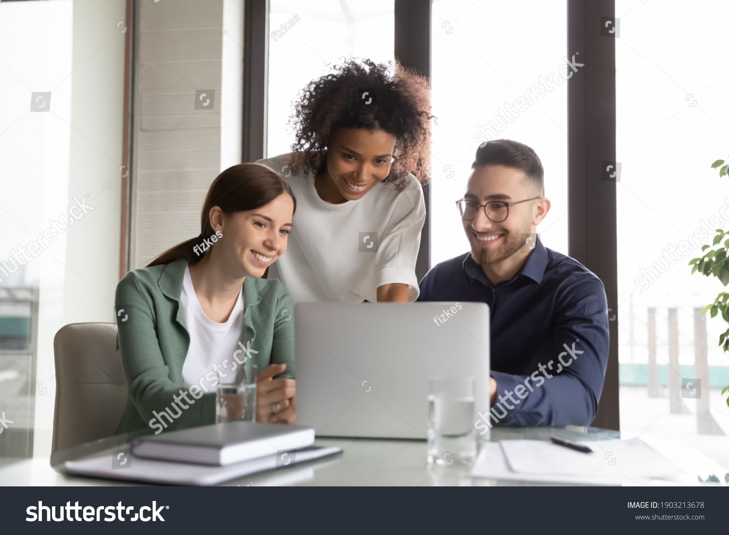 Three happy diverse colleagues working on project together, using laptop, sitting at table in office, discussing strategy, brainstorming, smiling African American businesswoman training staff #1903213678