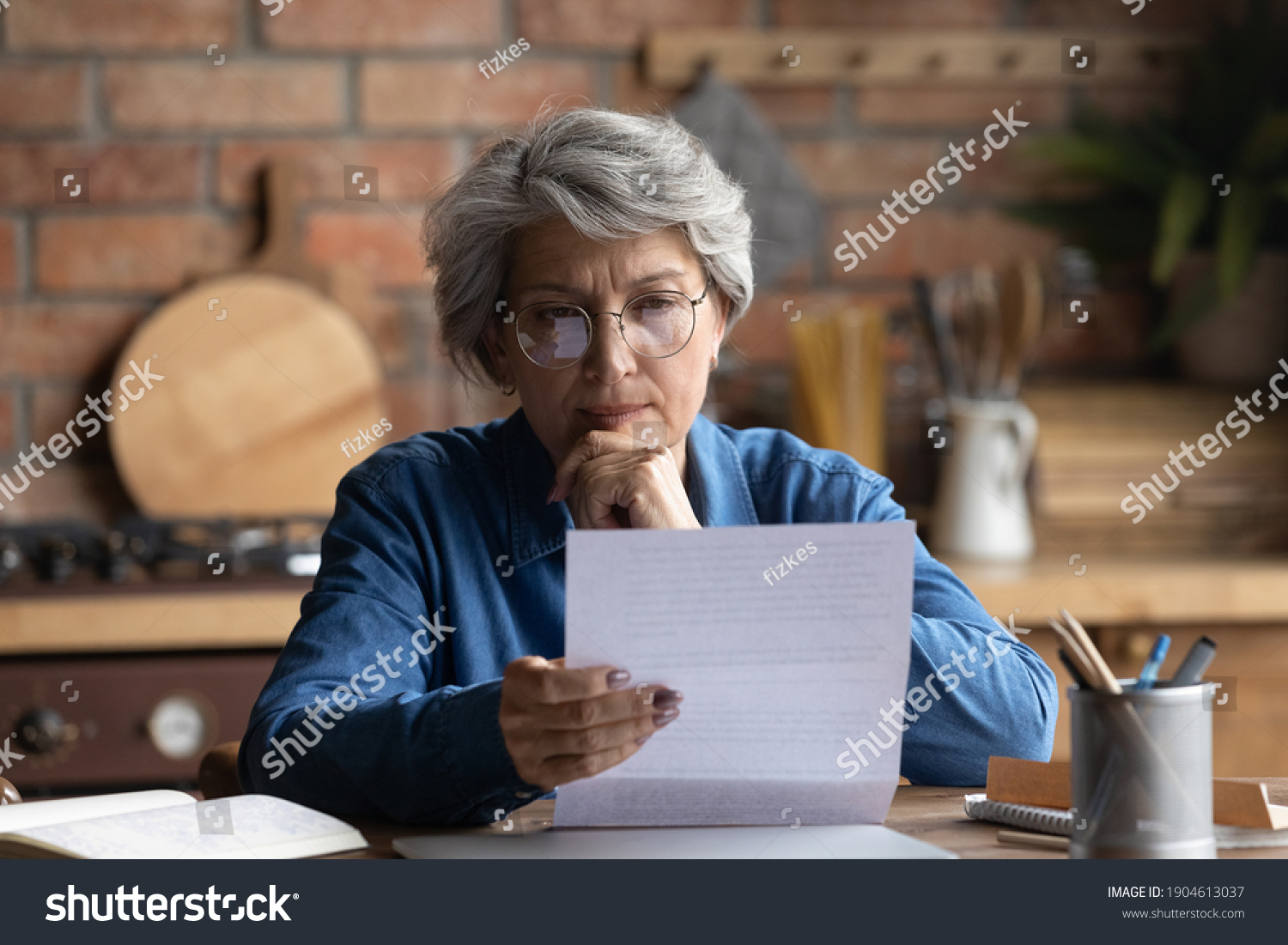 Thoughtful middle aged 60s woman in eyeglasses reading paper letter carefully. Pensive focused older mature retired lady looking at correspondence sheet. considering bank notification at home. #1904613037